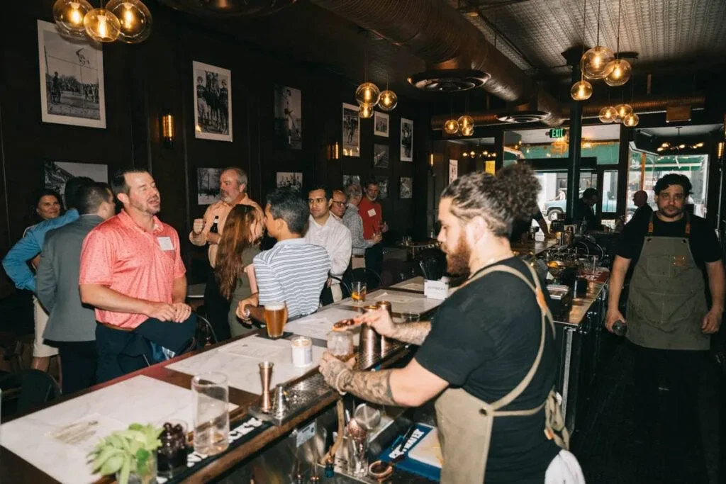 Busy bar interior: patrons, working bartenders, dark wood, warm lighting.