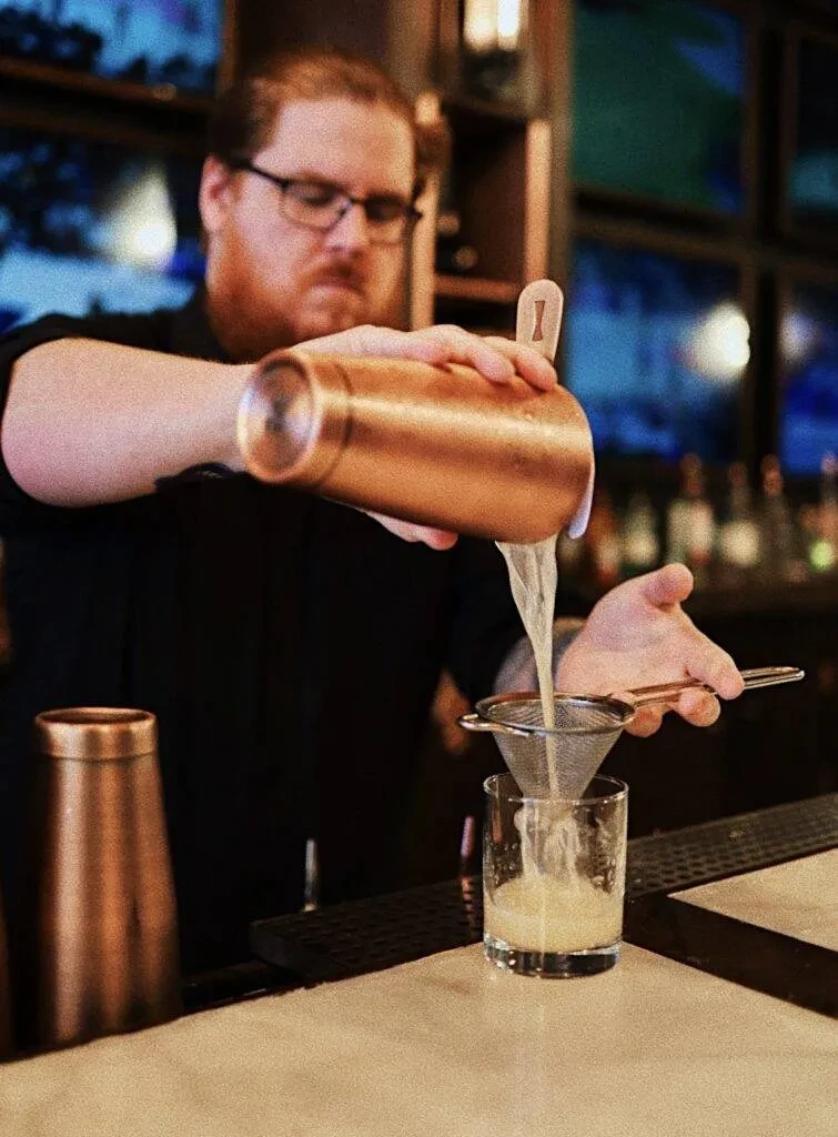 Bartender straining light liquid from copper shaker into a glass.