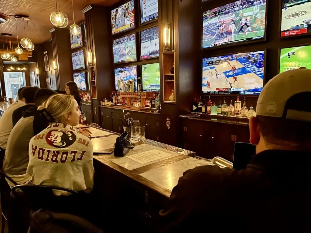 Sports bar interior with multiple TVs displaying sports, patrons at the bar.