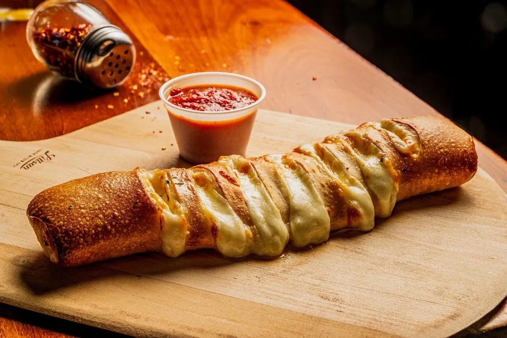 Golden-brown cheesy breadstick, oozing cheese, with dipping sauce on wood.