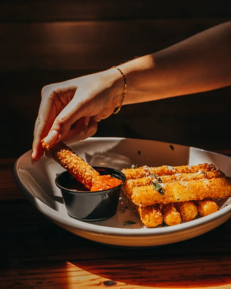Hand dips golden mozzarella stick into marinara sauce on a rustic table.