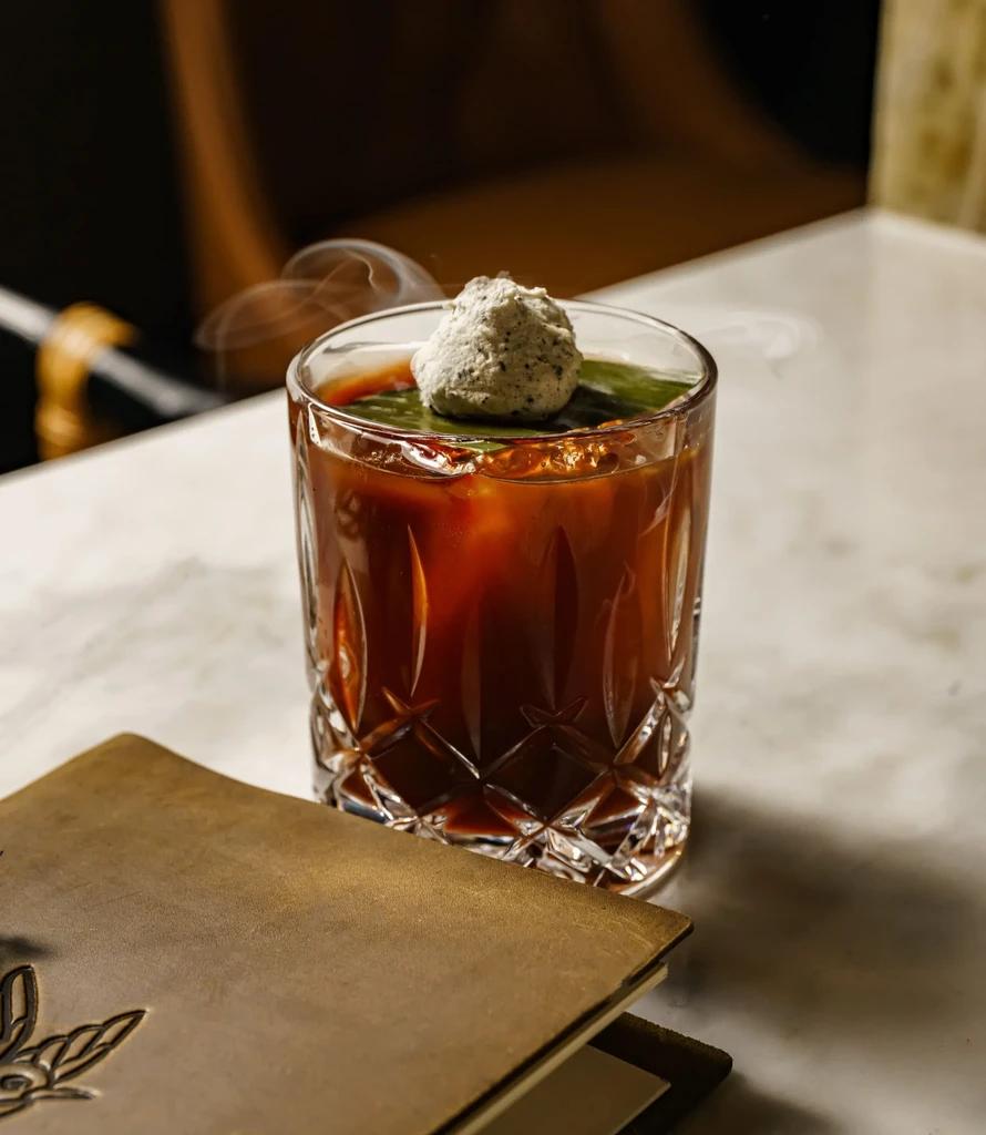 Dark cocktail in cut crystal glass with smoking spherical garnish and leaf.
