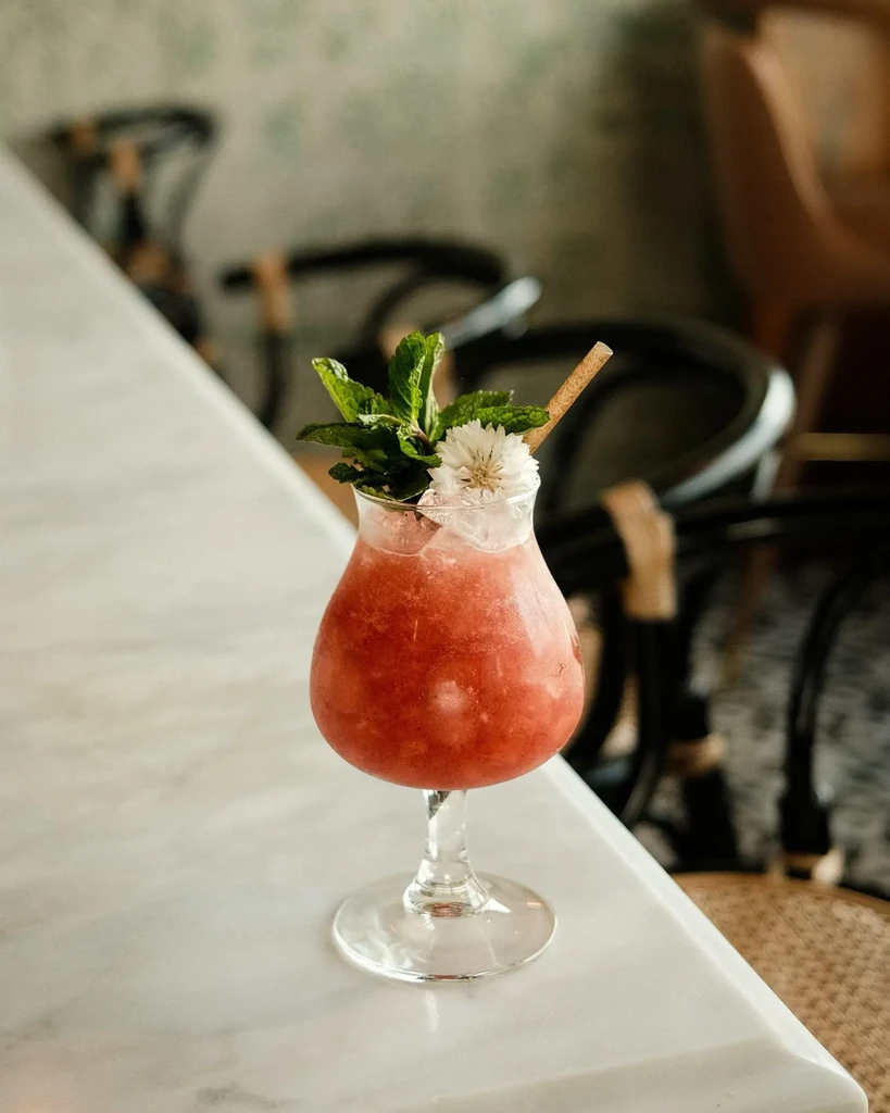 Reddish-pink frozen drink with mint, white flower, cinnamon garnish.