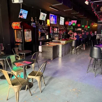 Dimly lit bar interior: patrons, stocked bar, TVs, and claw machines.