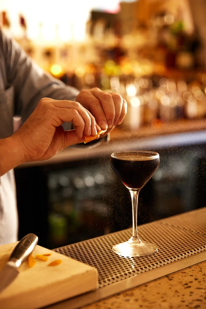 Hands squeezing orange peel to garnish a dark martini cocktail.