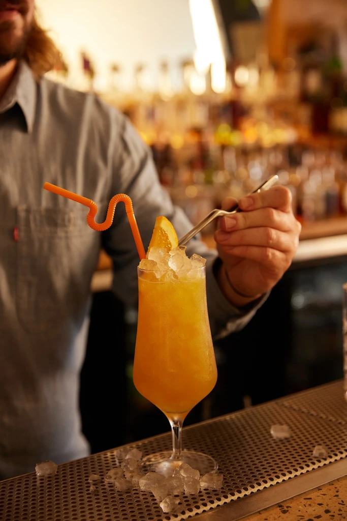 Orange cocktail with ice and straw, stirred by a bartender.