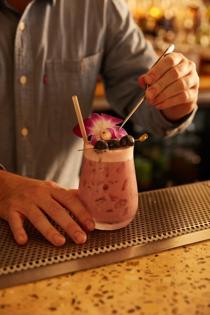 Close-up: bartender carefully garnishes pink cocktail with berries and an orchid.