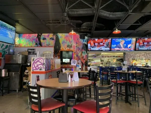 Restaurant interior: empty tables, colorful graffiti murals, bar, and TVs.