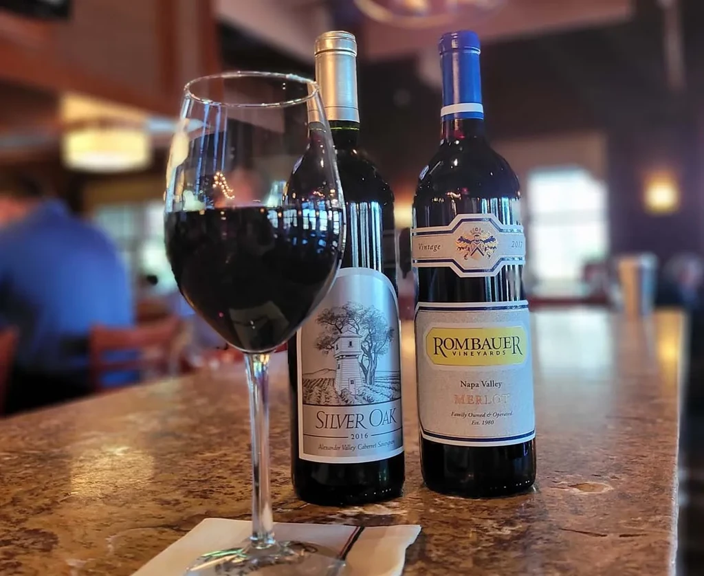 Wine glass, Silver Oak and Rombauer red wine bottles on a bar.