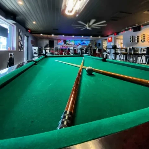 Green pool table with cues in a dimly lit bar, TVs in background.