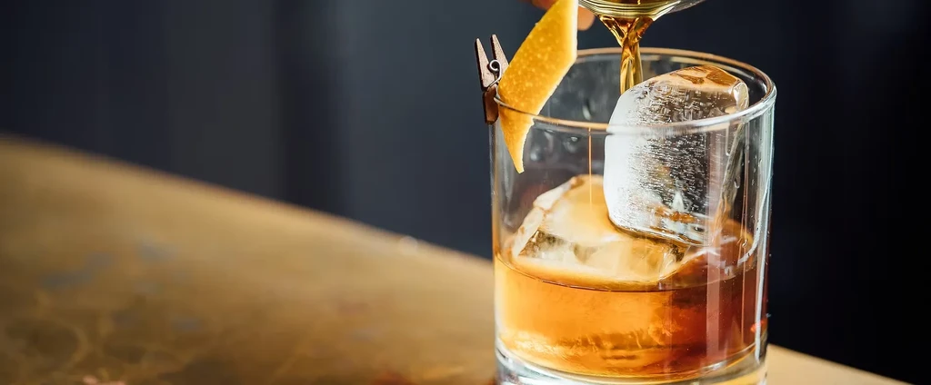 Amber liquid pouring into a glass with ice and lemon peel.
