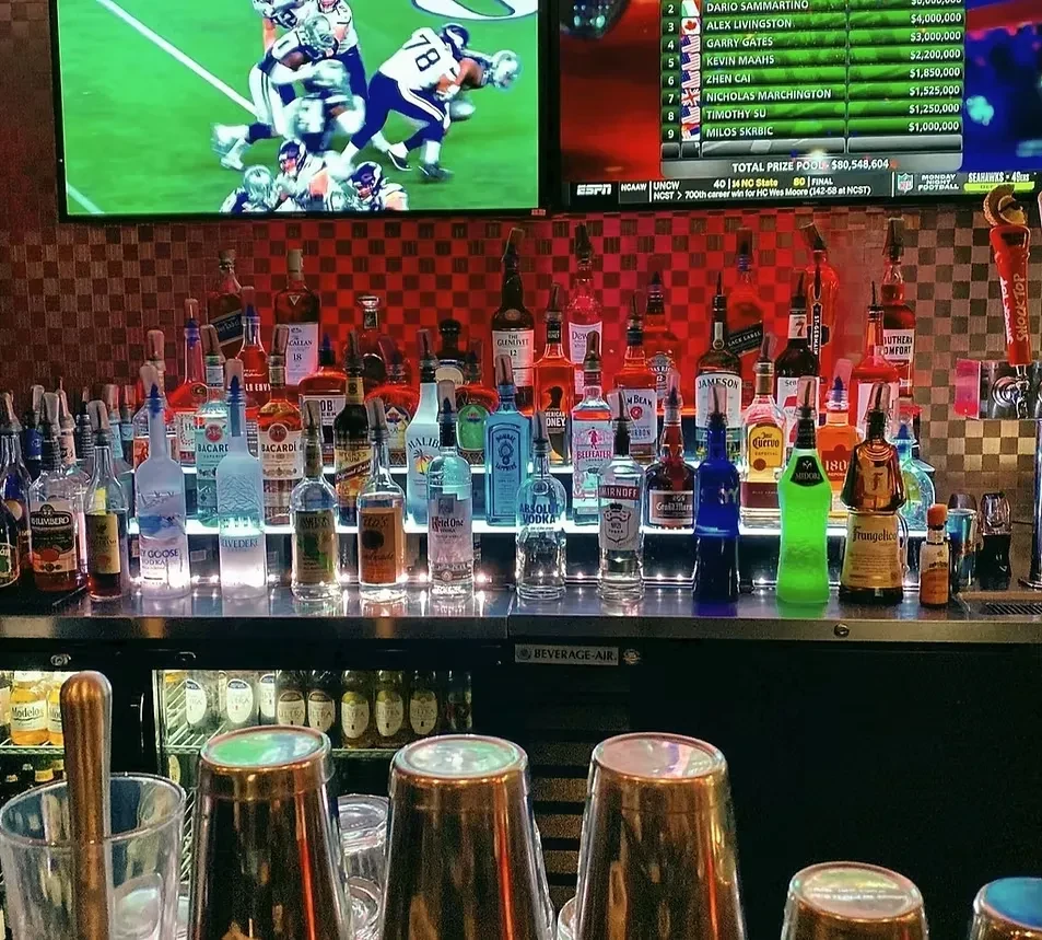 Illuminated bar counter with liquor bottles and two screens showing sports.