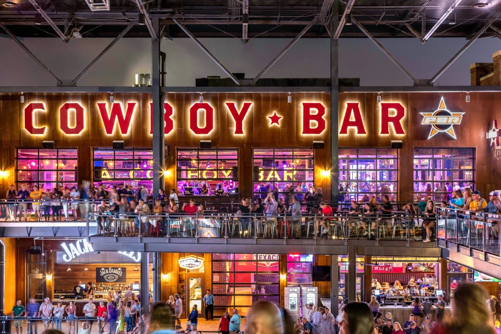 Illuminated "COWBOY BAR" sign on a crowded, multi-level bar at night.