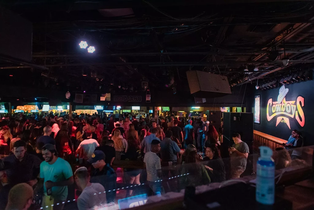 Crowded bar with colorful lighting and a "Cowboys" neon sign.