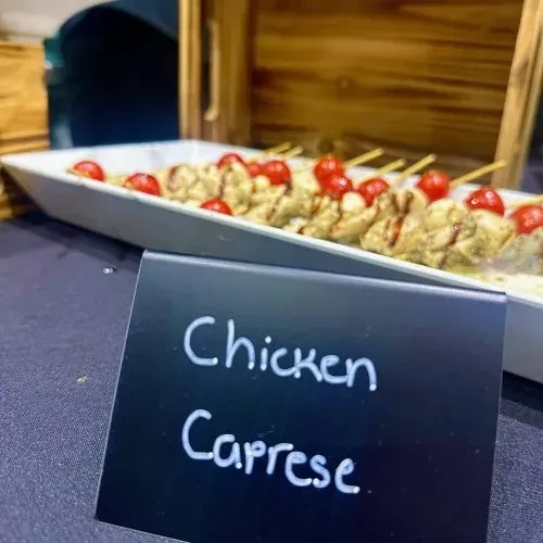 Close-up of Chicken Caprese skewers on a white platter with a sign.