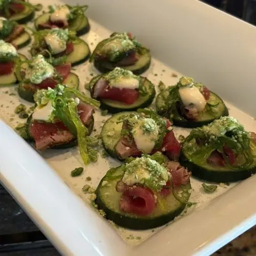 Platter of cucumber and seared tuna bites with creamy sauce and greens.