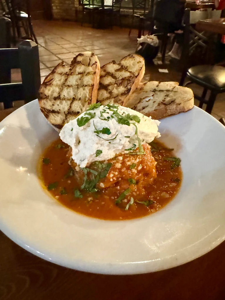 Rustic dish in bowl with red sauce, white cheese, herbs, grilled bread.