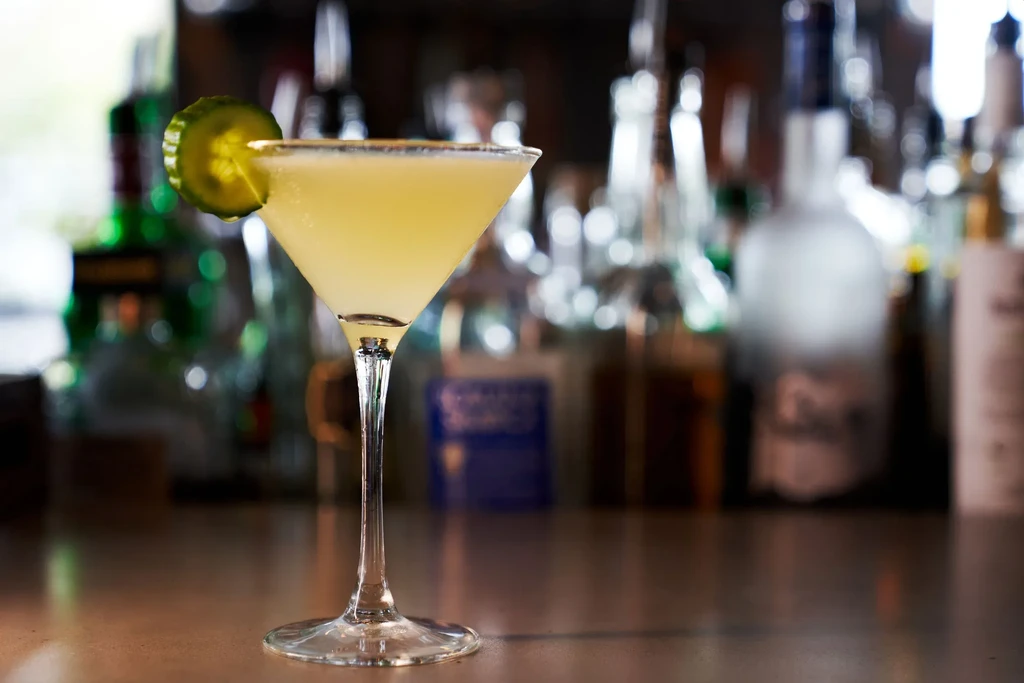 Pale yellow cocktail in a martini glass with cucumber and salted rim.