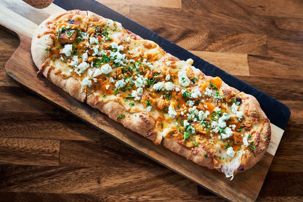Oval flatbread pizza with chicken, blue cheese, spicy sauce on wood.