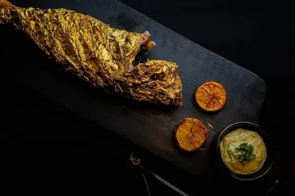 Whole fish covered in edible gold leaf, with citrus slices and sauce.