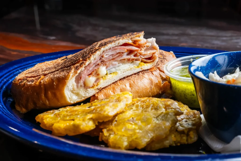 Cuban sandwich with tostones, green sauce, and white condiment on blue plate.