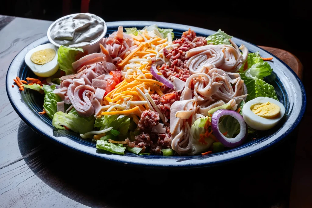 Large colorful chef salad with ham, turkey, cheese, eggs, and bacon.