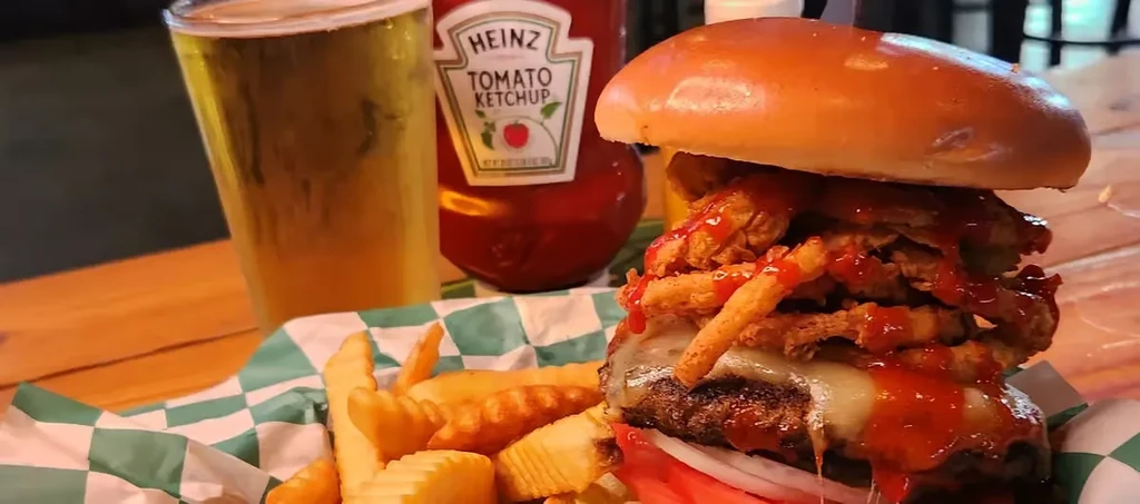 Stacked burger with onion rings, crinkle fries, beer, and ketchup.