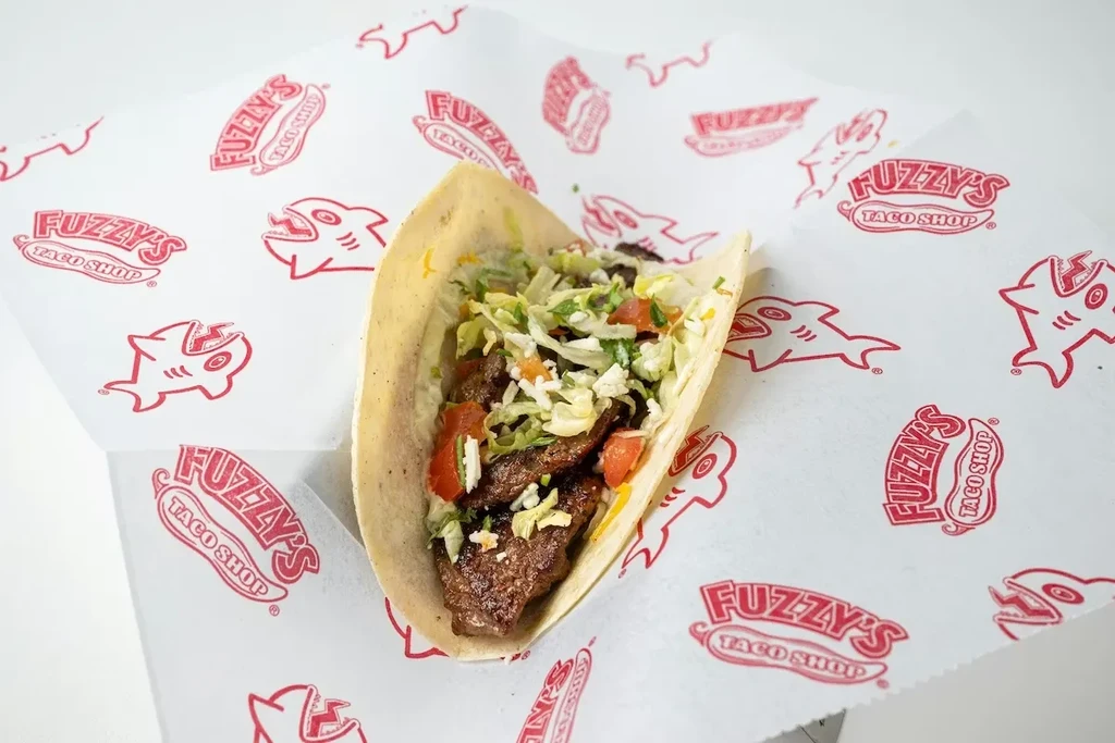 Grilled meat taco with lettuce, tomatoes, cheese on Fuzzy's logo liner.