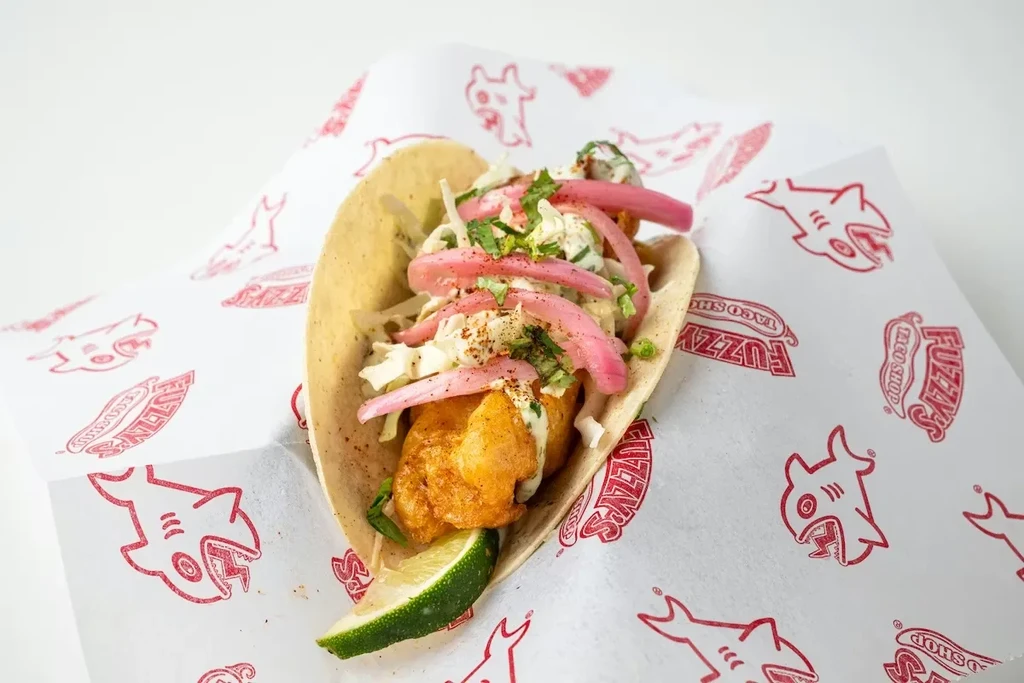 Single fried fish taco with slaw, sauce, pickled onions, and lime.