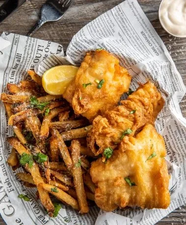 Golden-brown fish and chips on newspaper with lemon and herbs.