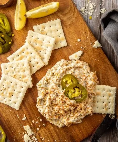 Creamy spiced dip with jalapeños, crackers, and lemon on a wooden board.
