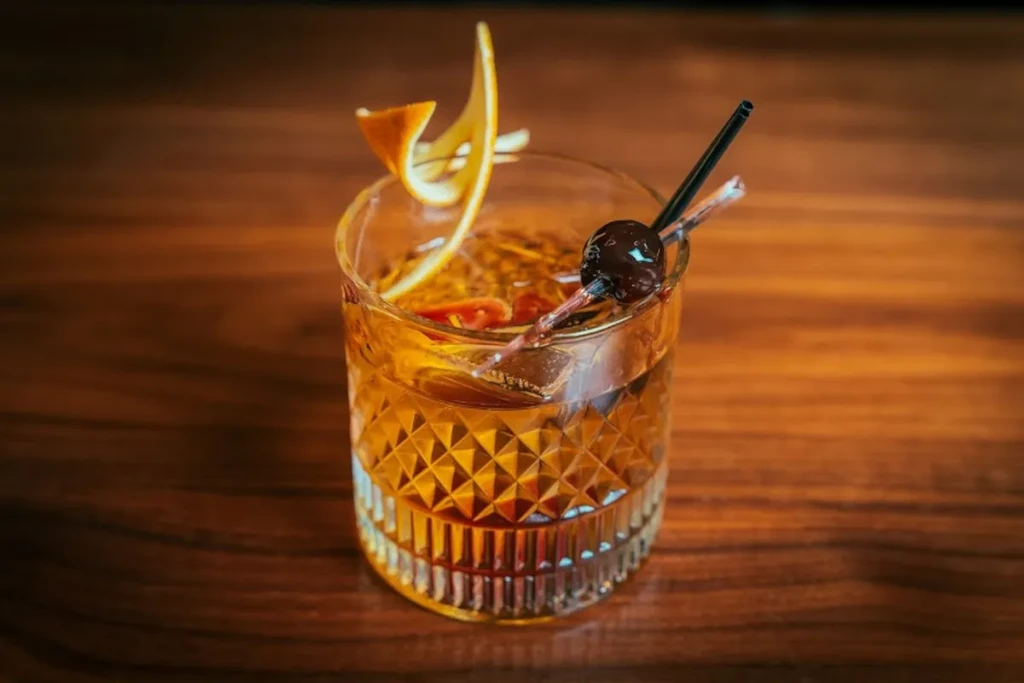 Amber cocktail in textured Old Fashioned glass with orange peel and cherry.
