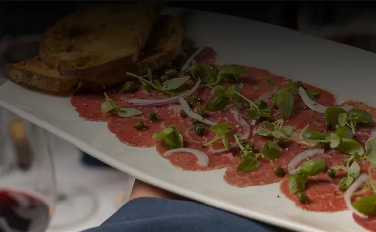 Plated beef carpaccio with herbs, red onion, capers, cheese, and toast.