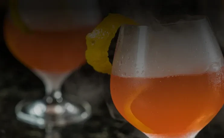 Two orange cocktails with citrus and smoke effect in a dark setting.