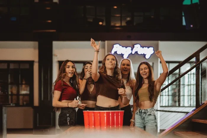 Five young women cheering at a beer pong game in a bar.