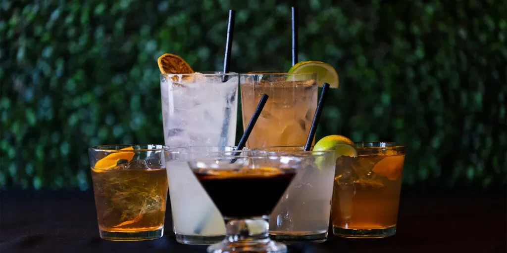 Seven diverse cocktails with garnishes on a dark surface and plant background.
