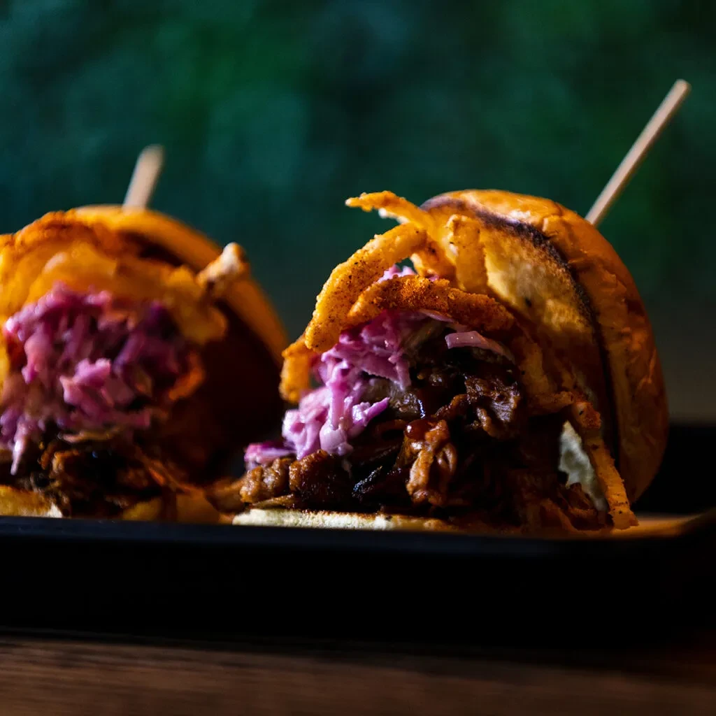 Gourmet pulled meat sliders with coleslaw and crispy onions on a dark dish.