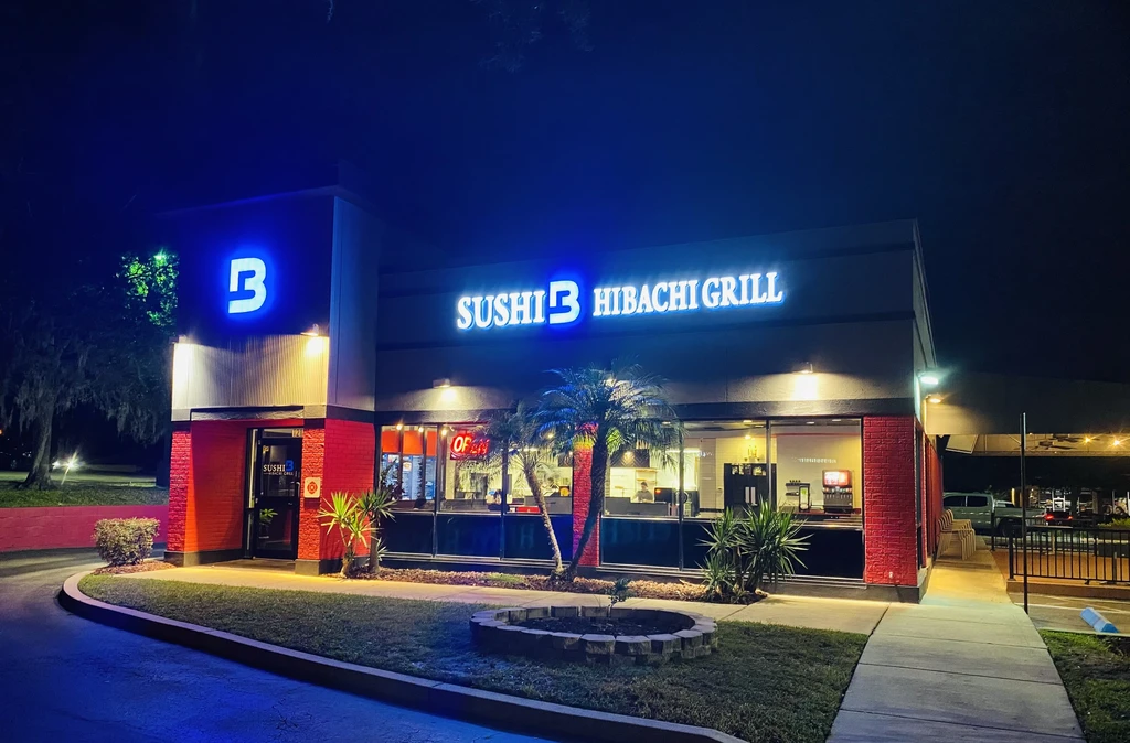 Nighttime exterior of Sushi B Hibachi Grill with neon signs and palm trees.