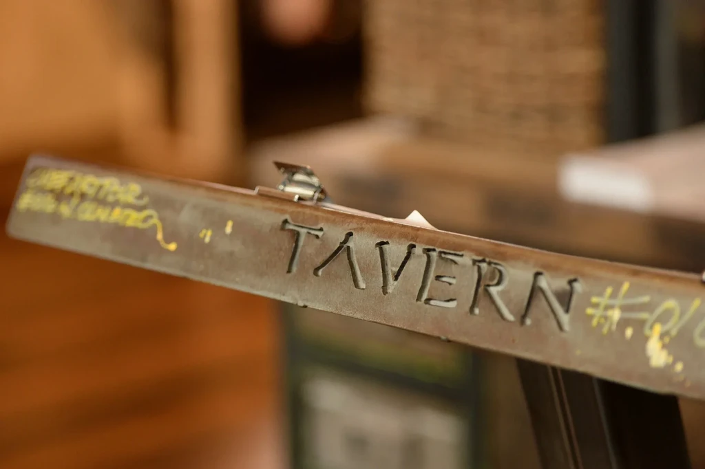 Rustic metal sign with "TAVERN" carved, yellow markings, and a metal clip.