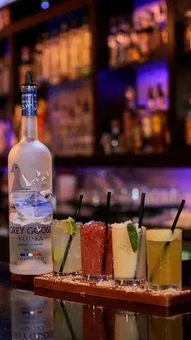 Grey Goose vodka bottle next to a four-cocktail flight on a bar.