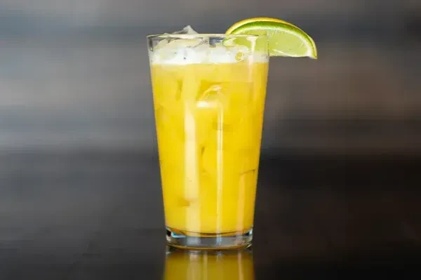 Tall glass of yellow-orange beverage with ice, lime, and froth.
