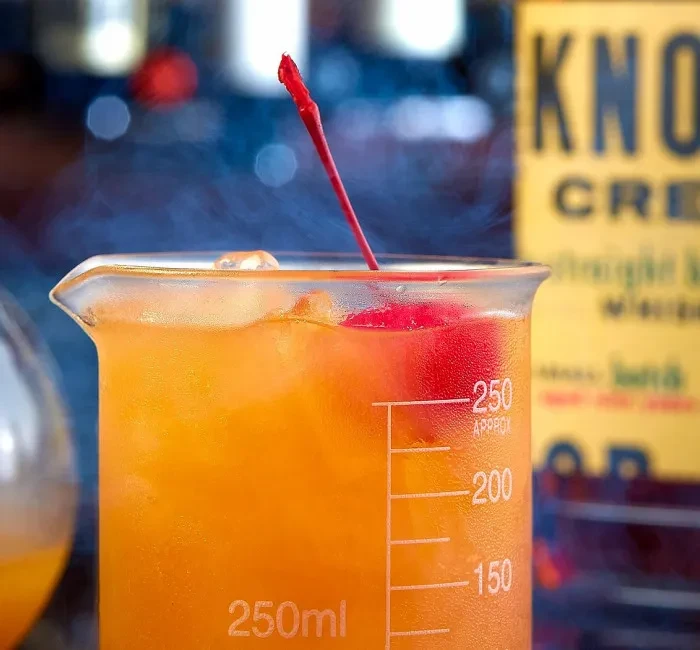 Vibrant orange cocktail in a lab beaker with cherry and mist.