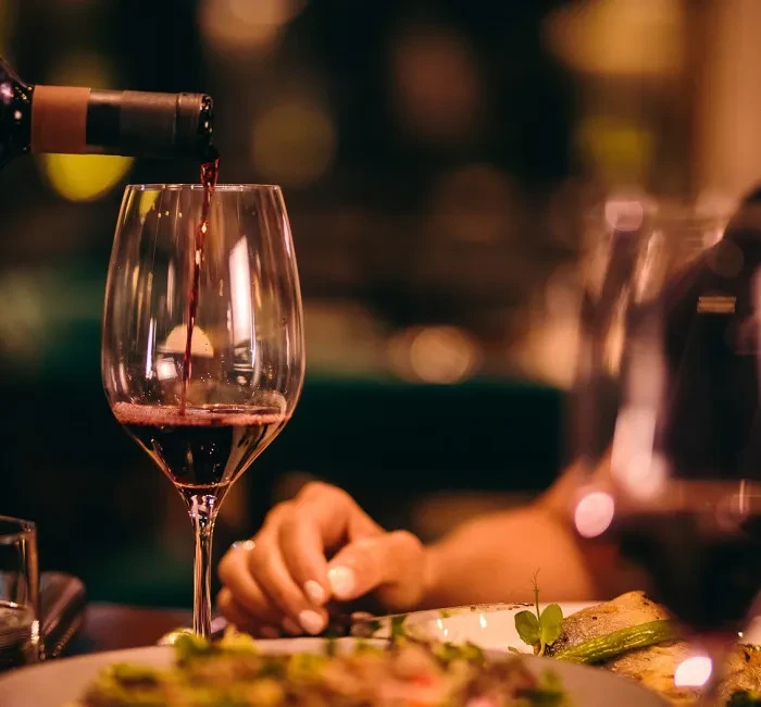 Red wine pours into glass; hand near food, warm blurred background.