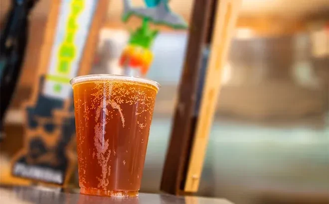 Amber carbonated drink in clear plastic cup, blurred colorful background.