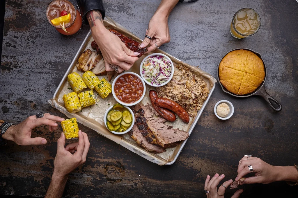 Communal barbecue platter with brisket, ribs, corn, and people's hands.