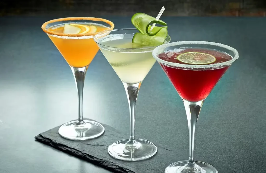 Orange, pale green, and red cocktails in martini glasses on a bar.