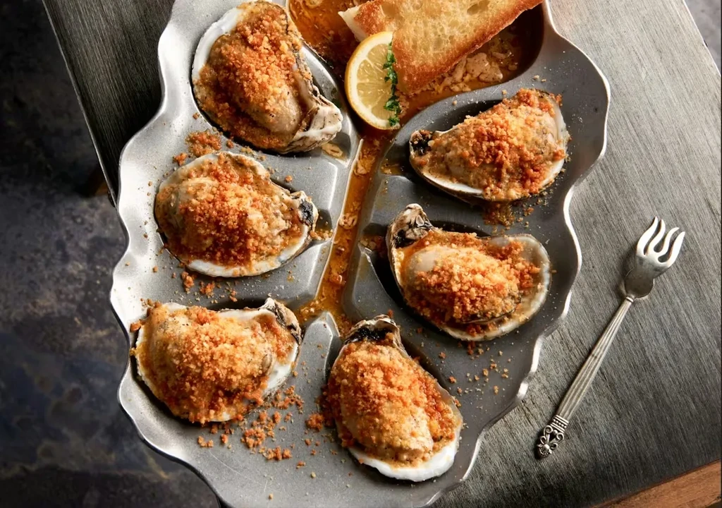 Six broiled oysters with breadcrumbs, lemon, and toast on a silver platter.
