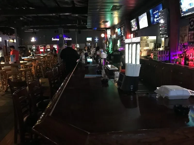 Dark bar interior with 'Backstage Billiards' sign, liquor bottles, and TVs.