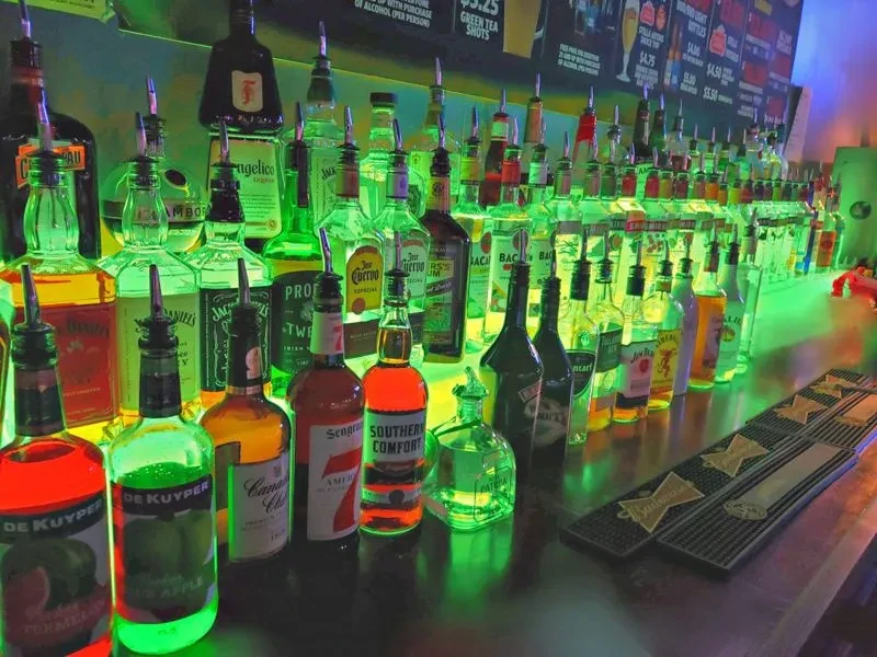 Bar counter with liquor bottles, illuminated by green and purple neon.