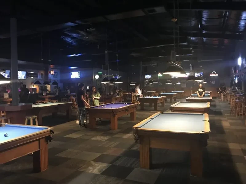 Dimly lit pool hall with many tables, a bar, and patrons.